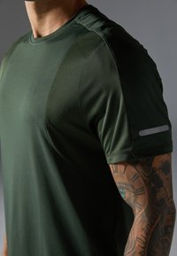 Green athletic t-shirt featuring smooth fabric with textured shoulder panels, round neckline, and reflective detail on the sleeve.