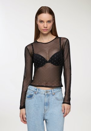 Top lungo a maniche trasparenti nere con un motivo a pois, dotato di outline di reggiseno nero e scollatura tonda. Abbinato a jeans in denim blu chiaro.