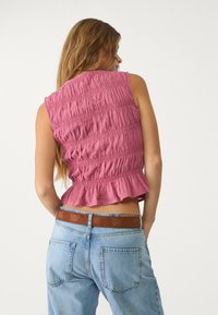 Rosa ärmlös topp med smockdetaljer, volang nederkant, i kombination med ljus denimjeans och ett brunt broderat bälte.