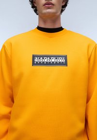 Geel sweatshirt van textuurstof met een zwart rechthoekig logo in het midden met de tekst "NAPAPIJRI geographic". Ronde hals.