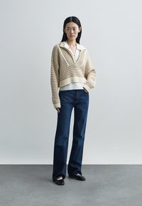 Μπεζ πλεκτό κοντό πουλόβερ με φερμουάρ και γιακά, φορεμένο πάνω από λευκό πουκάμισο και συνδυασμένο με σκούρο μπλε τζιν με καμπάνα και μαύρα επίπεδα παπούτσια.