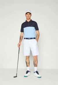 Αντρική γκολφ ενδυμασία που περιλαμβάνει ένα ναυτικό μπλε και ανοιχτό μπλε πόλο, λευκά σορτς και ένα ταιριαστό λευκό καπέλο. Εξοπλισμένος με μια γκολφ ρόπαλο, παρουσιάζει τα τατουάζ του.