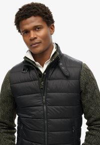 Gepolsterte schwarze Weste mit Reißverschluss, ausgestattet mit einem hohen Kragen und Details an den Schulterriemen, über einem strukturierten dunkelgrünen Pullover getragen.