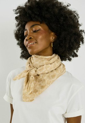 Mujer con cabello rizado que lleva pendientes de aro dorados, una bufanda beige con patrones de conchas atada alrededor del cuello y una camisa blanca con las mangas remangadas.