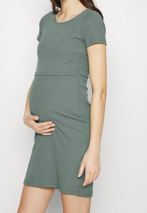 Mujer embarazada con vestido verde ajustado de manga corta, sosteniendo la barriga con una mano, vista desde los hombros hasta la mitad del muslo.