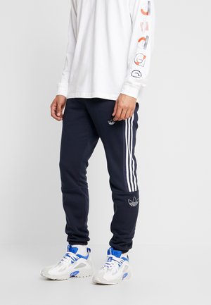 Osoba nosząca granatowe spodnie dresowe Adidas z białymi trzema paskami, białe sneakersy z niebieskimi akcentami oraz białą koszulę z długim rękawem z grafiką na rękawie.