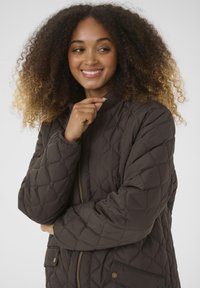 Veste en doudoune marron matelassée avec fermeture éclair à l'avant, col côtelé et poches latérales avec accents métalliques. Texture lisse et motif en losanges.