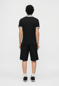 Άνδρας όρθιος, γυρισμένος με την πλάτη, φοράει μαύρο πόλο με λευκό γιακά και μανσέτες που έχουν κείμενο, μαύρο σορτς, λευκές κάλτσες και μαύρα αθλητικά παπούτσια.