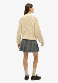 Beige kabelstrikket sweater med diamantmønstre, parret med en sort-hvid ternet plisseret nederdel. Sorte sko og lyse strikkede sokker fuldender looket.