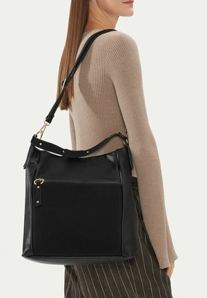 Donna che indossa un maglione beige a coste e una gonna a righe sottili, con una grande borsa a tracolla in pelle nera, con tasca frontale e dettagli in oro.