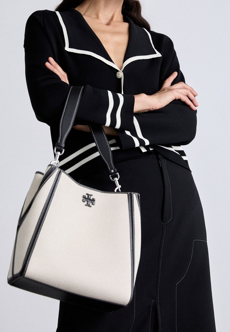 Personne portant une tenue noire avec un liseré blanc et tenant un sac à main blanc avec des bords noirs et des poignées noires doubles.