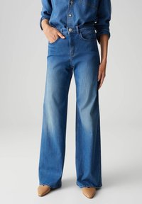 Jean en denim bleu à jambes larges, avec une taille haute, un léger délavage près des cuisses et des poches robustes. Finition avec une fermeture à bouton et zip.