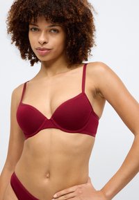 Βουργουνδί μπουστάκι με λεία, μαλακά cups και ρυθμιζόμενους ιμάντες. Κατασκευασμένο από ελαστικό βαμβάκι. Περιλαμβάνει λεπτομέρεια λογότυπου στους ιμάντες.