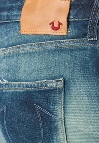 Jeans de mezclilla con un lavado azul desgastado. Parche de cuero natural cosido en la cinturilla, con un logo rojo. Costuras en contraste en los bolsillos.