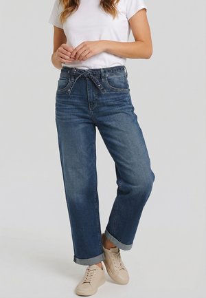 Vrouw draagt opgerolde blauwe jeans met een gebonden riem, wit t-shirt en beige sneakers, staand met ontspannen handen voor zich.