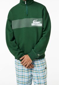 Sweat à capuche vert à fermeture éclair avec un motif à carreaux blancs et un logo sur la poitrine. Comprend des poignets et un ourlet côtelés, associé à un pantalon à carreaux.