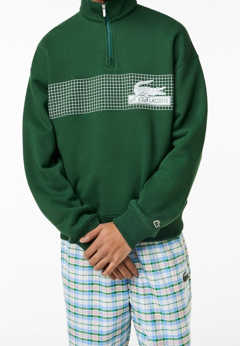 Sweat à capuche vert à fermeture éclair avec un motif à carreaux blancs et un logo sur la poitrine. Comprend des poignets et un ourlet côtelés, associé à un pantalon à carreaux.