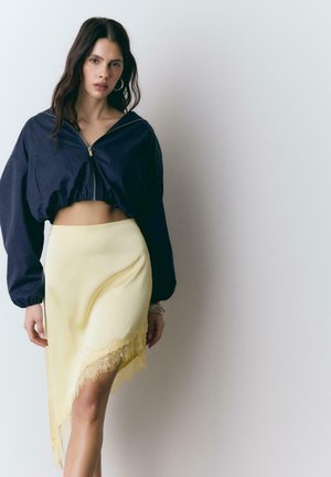 Donna che indossa una giacca corta con zip blu navy e una gonna asimmetrica color giallo pallido con rifiniture in pizzo, in piedi davanti a uno sfondo chiaro e uniforme.
