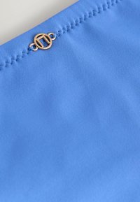 Bolsa de tela azul con costuras visibles y un acento de hardware dorado circular que presenta iniciales entrelazadas. Textura suave.