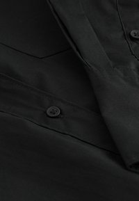 Gros plan sur un tissu noir avec un bouton et une partie de manche, montrant les détails de couture et de couture.