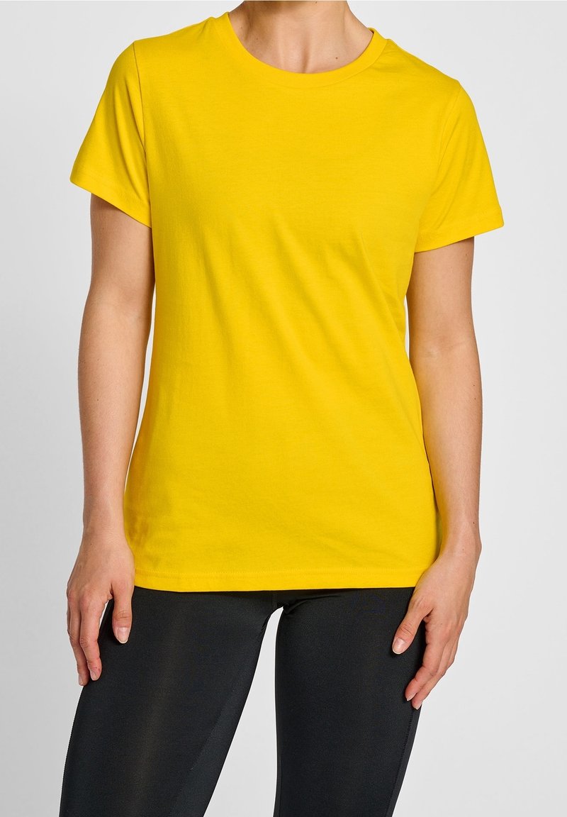 Personne portant un t-shirt jaune uni à manches courtes et un pantalon noir, debout avec les bras naturellement pendus le long du corps.