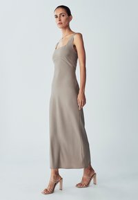 Maxikleid aus weichem beigen Stoff mit quadratischem Ausschnitt und dünnen Trägern. Kombiniert mit hellbeigen High-Heels. Glatte Textur.
