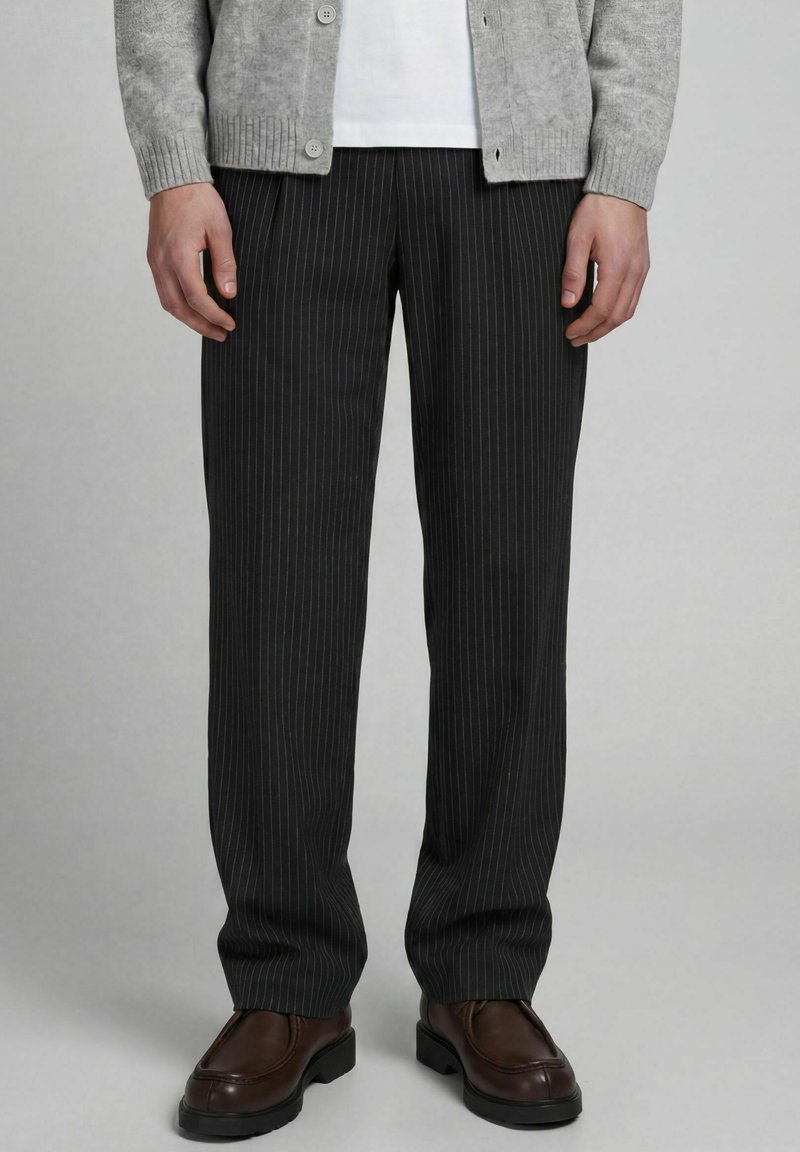 Homme portant un pantalon noir à fines rayures, des chaussures en cuir marron, un cardigan gris boutonné et une chemise blanche, debout sur un fond gris.