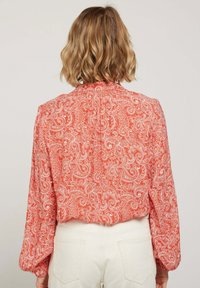 Blouse à motif paisley rouge avec des manches longues, un empiècement au dos froncé et une coupe légèrement cropped, associée à un pantalon taille haute de couleur claire.