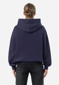 Navyblauer Hoodie mit großer Kapuze, lockerer Passform und gerippten Bündchen; getragen über dunkelgrauen Jeans; weicher Stoff mit minimalen Details.
