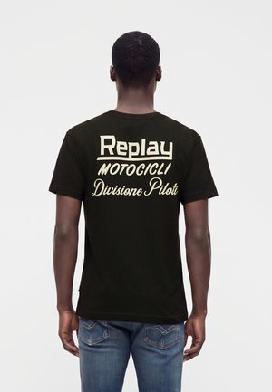 Homme portant un t-shirt noir avec texte blanc "Replay Motocicli Divisione Piloti" dans le dos et un jean bleu, debout de dos.