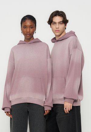 Two young adults wearing oversized dusty pink hoodies and dark pants, standing side by side against a plain background.