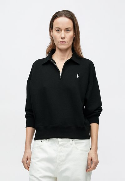 Svart hettejakke med glidelås og krage, med subtil logo på brystet. Ribbestrikkede mansjetter og kant gir en avslappet passform. Brukt med hvite bukser.