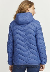 Doudoune bleue avec un motif en chevrons, dotée d'une capuche, d'une texture matelassée et de poignets élastiques. Le tissu a l'air lisse et isolant.
