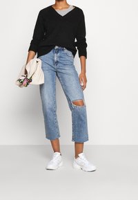 Schwarzer V-Ausschnitt-Pullover kombiniert mit hellblauen, knöchellangen Jeans mit einem aufgeschlitzten Knie. Das Model hält eine strukturierte, hellfarbige Tasche und trägt weiße Sneaker.