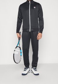 Svart idrottsoverall med vit dragkedja, med en logotyp på vänster bröst, kombinerad med vita och marinblå tennisskor och håller en tennistennis.