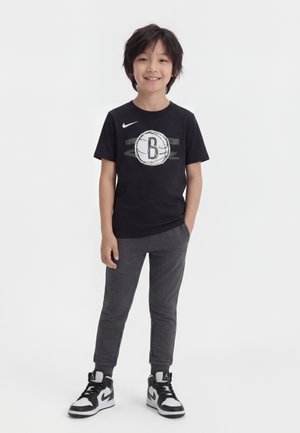 Garçon souriant aux cheveux noirs portant un t-shirt noir des Brooklyn Nets, un pantalon de survêtement gris foncé et des baskets noir et blanc, debout sur fond blanc.