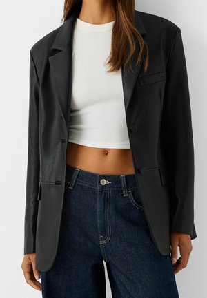 Frau trägt schwarzen Lederblazer über weißem, kurz geschnittenem, geripptem Top und hoch taillierter dunkelblauer Jeans, steht vor einfachem Hintergrund.