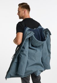 Giacca parka impermeabile blu con cappuccio, dotata di una morbida fodera in pile e di diverse tasche. Indossata a spalla con una maglietta nera.
