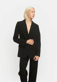 Mustaherra blazer, jossa on lovetut läpät, yksi tasku ja napilliset hihat. Puettu mustan yläosan päälle ja yhdistetty samansävyisiin housuihin. Sujuva tekstuuri.