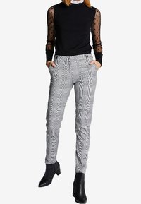 Haut long à manches en mesh noir associé à un pantalon tailleur à carreaux gris. Le pantalon présente une coupe slim et un ourlet roulé, complété par des bottines à talons noirs.