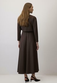 Rochie lungă, maro, dintr-un material moale, cu un jachetă scurtă și mâneci lungi. Prezintă o fustă fluidă și sandale negre cu vârf deschis.