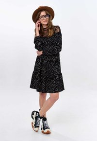 Zwarte jurkje met stippen, lange mouwen en kantafwerking, gelaagde zoom, gecombineerd met zwarte en crèmekleurige sneakers en een bruine hoed met brede rand.