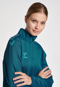 Maglia sportiva zip-up color turchese con colletto alto, dotata di un logo e dettagli a strisce in turchese chiaro sulle spalle. Tessuto liscio ed elastico.