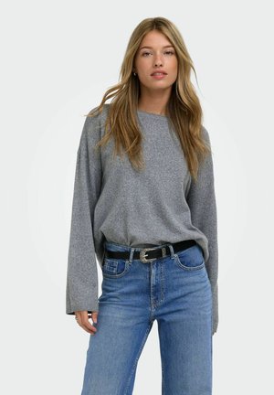 Femme aux longs cheveux blonds portant un pull gris ample et un jean bleu avec une ceinture noire, debout devant un fond clair uni.