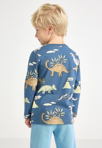 Sininen pitkähihaine paita, jossa on dinosauruksia ja pilviä keltaisen, vihreän ja valkoisen sävyissä. Pehmeää materiaalia ja pyöreä pääntie.