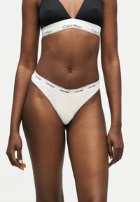 Γυναίκα που φοράει μαύρο μπραλέτι και λευκά εσώρουχα Calvin Klein με λογότυπο στη μέση, στέκεται μπροστά σε απλό φωτεινό φόντο.