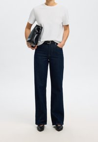 Personne portant une chemise blanche à manches courtes, un jean bleu foncé à jambes larges, une ceinture noire, des chaussures noires et tenant un sac à main noir sur le côté gauche.