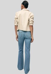Giacca beige corta con maniche arrotolate, abbinata a jeans azzurri svasati. Il denim ha un orlo grezzo e dettagli nelle tasche posteriori.