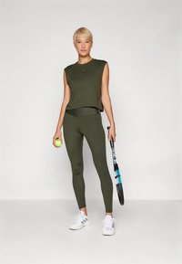 Mulher vestida com um conjunto atlético verde-oliva, segurando uma bola de ténis e uma raquete, usando sapatilhas brancas, em pé contra um fundo branco simples.