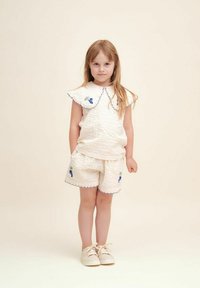 Hvid quilted top med bølget krave og blå blomsterbroderi, parret med matchende shorts. Let tekstur, afslappet pasform og beige sko.
